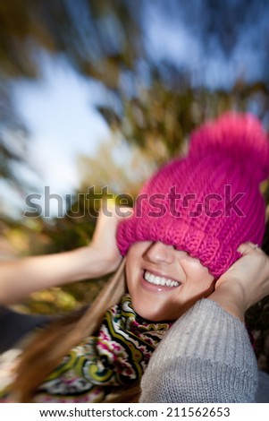 Color explosion in winter fashion with a playful woman pulling a vivid magenta knitted beanie hat over her eyes with a mischievous smile as she stands outside on a cold day