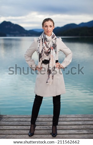 Proud beautiful stylish woman standing on a jetty posing confidently with her hands on her hips in an elegant coat and scarf