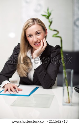 Businesswoman sitting at her desk writing notes looking at the camera with a thoughtful expression and friendly smile