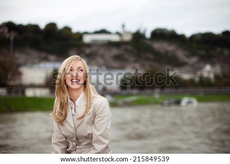 Happy city girl with long blond hair in a stylish coat standing laughing happily and looking into the air, with copy space