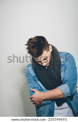 Sad Handsome Man Wearing Denim Jacket and Gray Knitted Scarf, Crossing his Arms in Front his Body While Looking Down. Isolated on Light Gray Background.