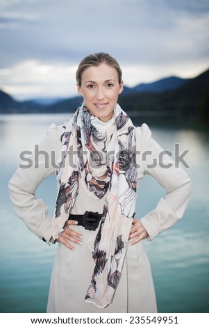 Confident stylish woman in a beige coat and elegant scarf standing with her hands on her hips at a mountain lake looking at the camera with a friendly smile