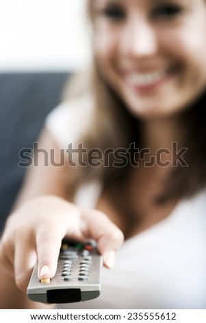 Smiling woman changing programs on a TV using a remote control aimed at the viewer, focus to the remote control