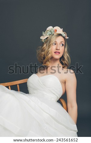 Relaxed young bride in her white bridal gown and veil with orchids in her hair reclining in a chair looking to the side with a serious expression, over grey