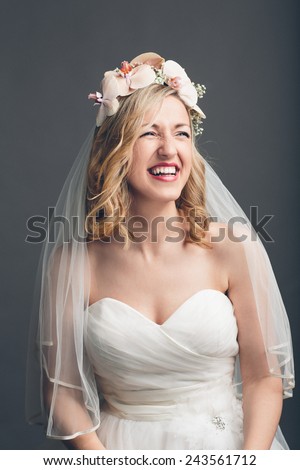 Charming vivacious young bride in a strapless white wedding dress and veil with flowers in her hair standing laughing, upper body over grey