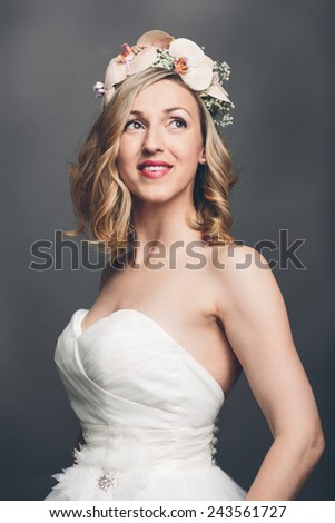 Elegant pretty bride with flowers in her hair wearing a strapless white bridal dress standing looking meditatively up into the air with a tender smile, upper body over grey