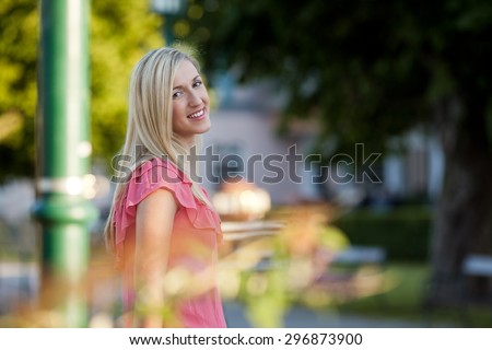 Half Body Shot of a Pretty Blond Young Woman Standing on the Road Side beside a Post and Smiling at the Camera.