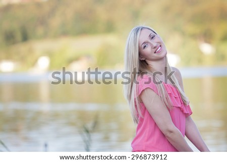 Half Body Shot of a Pretty Blond Young Woman Standing at the Lakeside and Smiling at the Camera
