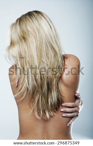 Close up Rear View of a Shirtless Blond Young Woman Hugging her Self While Looking on the Side Against Light Gray Wall Background.