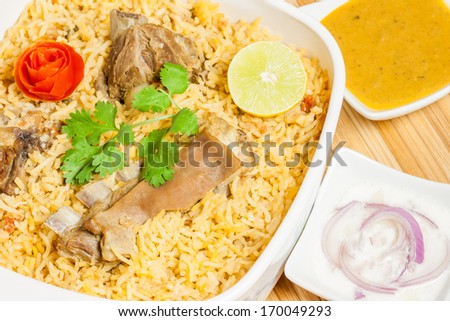 Mutton Biryani - Closeup view from the top of delicious mutton (lamb) biryani garnished with tomato peel, cilantro and lemon. It is served with onion salad (raita) and vegetable curry.