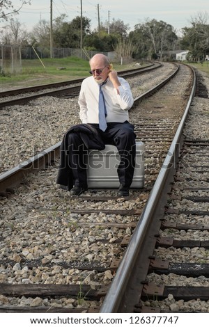 Jobless senior businessman sits on suitcase on railroad train tracks pondering his uncertain future, wiping his hot sweaty neck with a handkerchief.