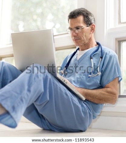 Mature doctor sitting on floor, leaning against wall, in blue scrubs, working on his laptop