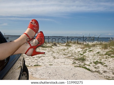 Woman\'s legs with red patent leather spike heels dangling out the car window parked at the beach