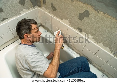Man working in a tight space while applying ceramic tile to a bathtub enclosure wall. Viewed looking down.