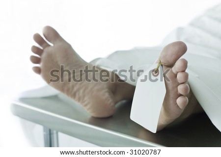 Photo of body covered with a sheet in a morgue, feet toward camera with blank tag on the big toe. Feet are clean and smooth. (body is a model)