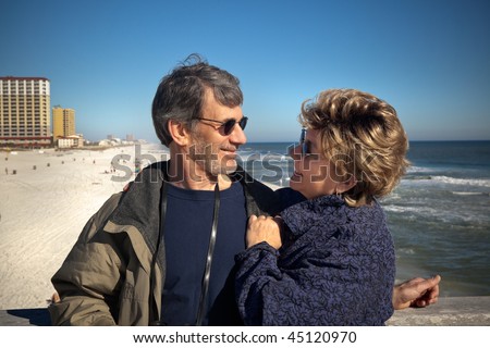 Happy senior couple enjoying themselves at a beautiful beach on their winter or spring vacation. Wide view of Couple gazing at each other with the beach, ocean and hotels in the background.