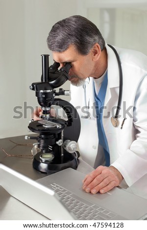 A male doctor or scientist looking through a microscope on a table with laptop computer.