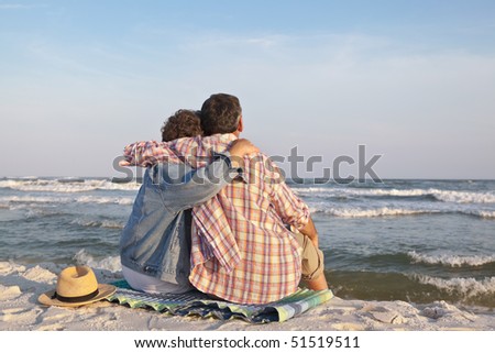 Affectionate mature couple sitting close together at sunset on the beach in the sand, gazing out to sea.