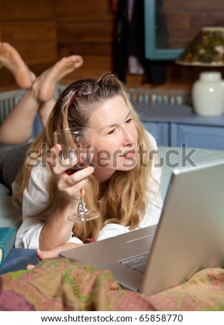 Attractive mature woman relaxing on a bed with glass of wine and laptop computer