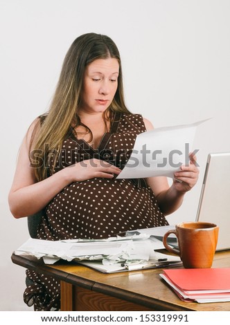 Pregnant mother-to-be reads bills and bank statements while seated at a desk with stacks of unpaid bills next to laptop computer. She looks surprised and slightly worried.