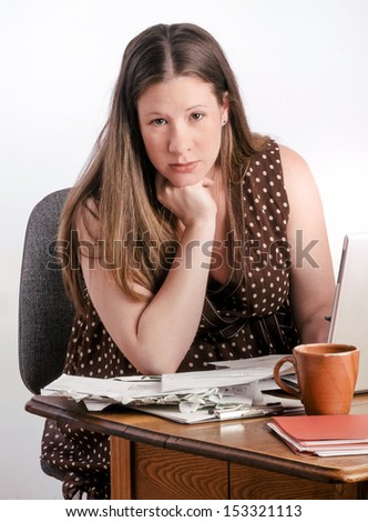 Pregnant mother-to-be reads bills and bank statements while seated at a desk with stacks of unpaid bills next to laptop computer. She looks calm and confident.