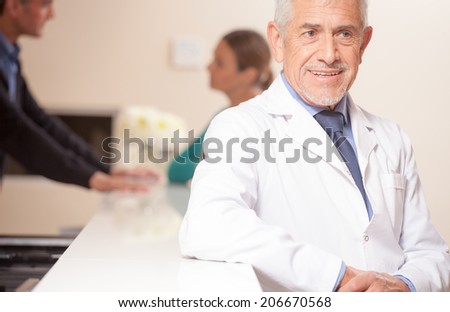 Smiling senior male doctor smiling at hospital reception desk, female patient speaking with young doctor on background.