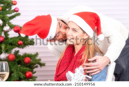 Happy Christmas family scene. Husband wearing red hat kissing wife under tree.