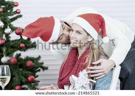 Happy Christmas family scene. Husband wearing red hat kissing wife under tree.