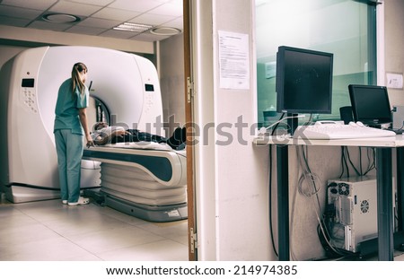 Hospital room with computer and monitors. Female doctor and male patient undergoing scan in adiacent room.