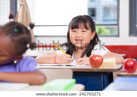 Asian girl smiling at classroom desk drawing on her book during a school lesson.