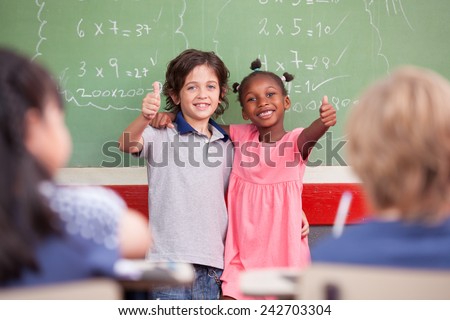 Caucasian kid and afro american female smiling to the classroom in front of chalkboard. Primary school concept.