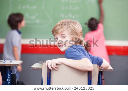 Multi ethnic elementary classroom. Kid looking at camera while classmates at chalkboard.