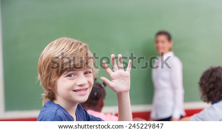 Kid raising hand in classroom with teacher explaining at chalkboard.