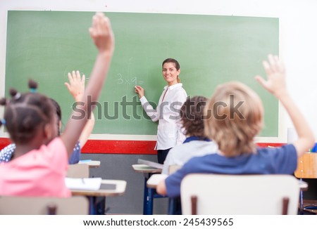 Intelligent group of young school children raising their hands in the air to answer a question posed by the female teacher, view from behind.
