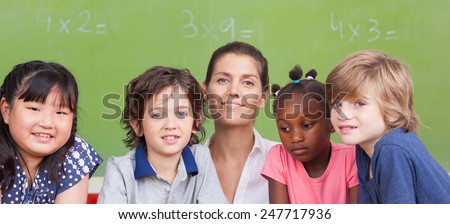 Multi ethnic elementary classroom with female teacher in front of green chalkboard.