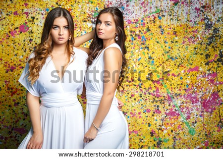Pretty twins girls in white long dresses posing over painted yellow background