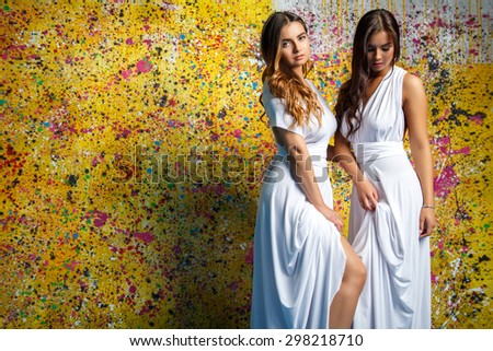 Pretty twins girls in white long dresses posing over painted yellow background