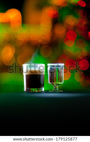 A glass of hot coffee standing next to a glass filled with liquor with colored bokeh lights in the background