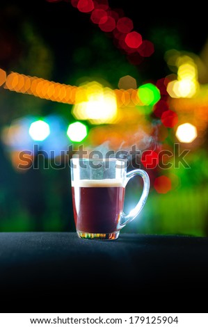 A glass mug filled with hot coffee standing on a table with colored bokeh lights in the background