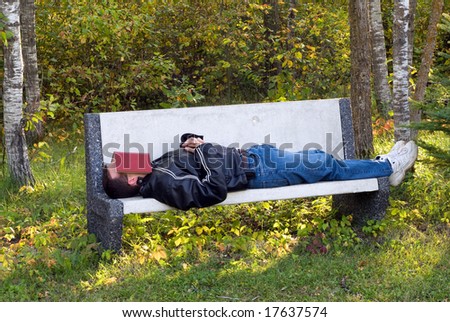 A young man sleeping on a park bench with a book over his face