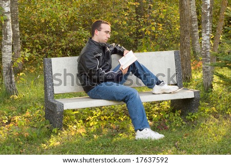 A young caucasian man sitting on a park bench reading outside