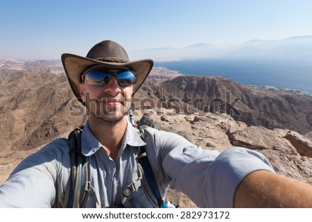 Smiling happy man hiker tourist wearing hat selfie picture on mountain summit above Red sea , Eilat city, Israel.