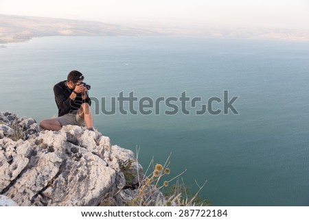 Man photographer sitting taking picture  resting stone cliff above lake sea, Arbel mountain, Kineret Galilee Tiberias lake, Israel.
