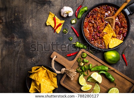 Chili con carne in frying pan on dark wooden background. Ingredients for making Chili con carne. Space for text. Top view. Chili with meat, nachos, lime, hot pepper. Mexican/Texas traditional dish
