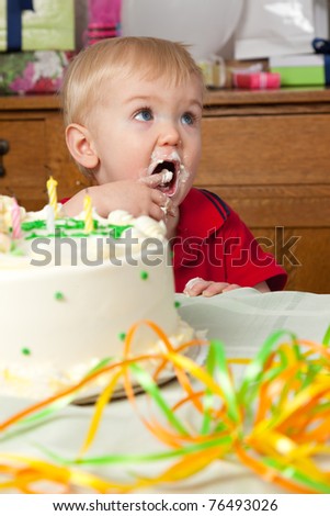 A toddler boy sneaks a taste of the icing on his birthday cake while looking up at his mom.