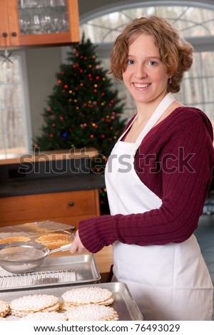 A young woman wearing a white apron sprinkles powdered sugar onto freshly baked pizzelles with Christmas Tree in background.