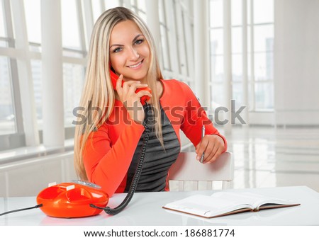 a young beautiful smiling caucasian businesswoman sitting at table speaking with phone and looking at camera