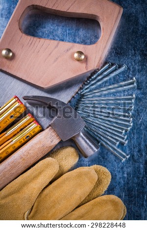 Hand saw protective gloves construction nails wooden meter and claw hammer on scratched metallic background close up view building concept.
