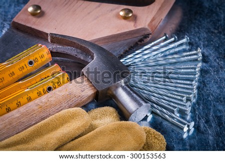 Hand saw leather gloves construction nails wooden meter and claw hammer on scratched metallic background top view building concept.