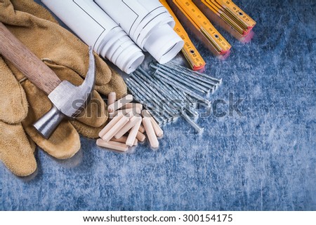 Blueprint rolls claw hammer safety gloves wooden meter metal nails and dowels on scratched metallic surface construction concept.